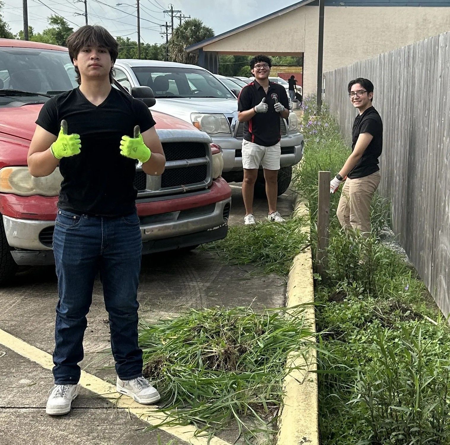 Three young men weeding by a fence. One gives thumbs up, wearing gloves, standing by parked trucks.