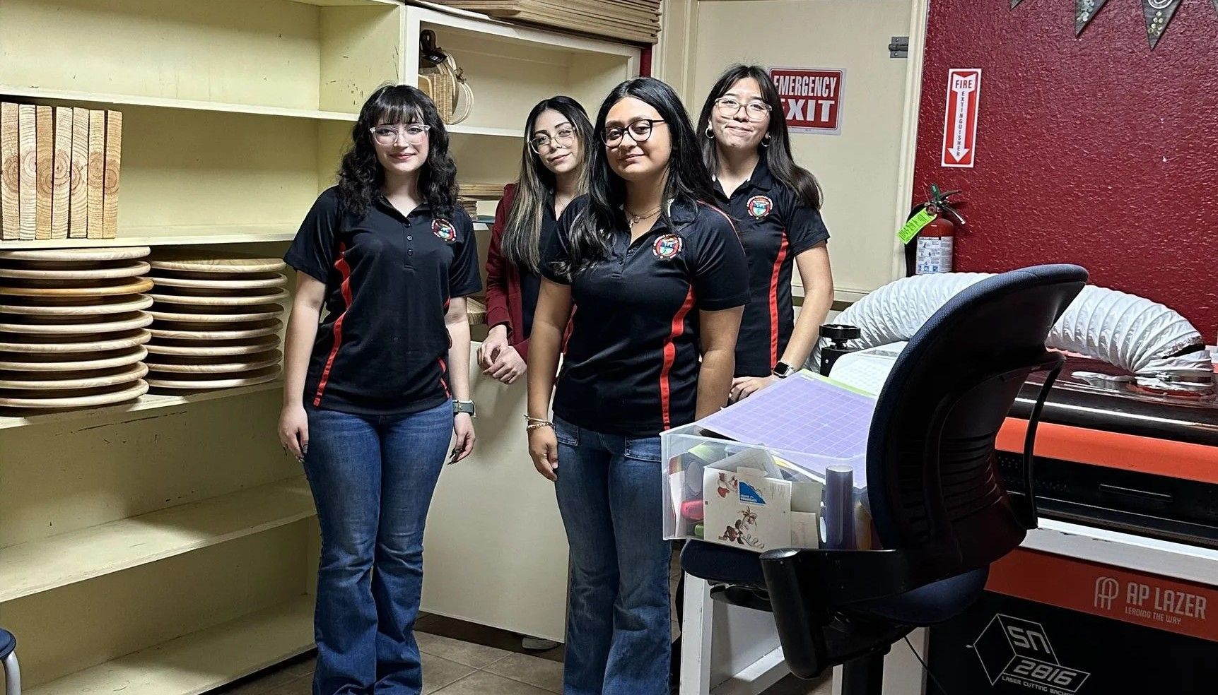 Four young women in black shirts and jeans pose next to a machine in a room.