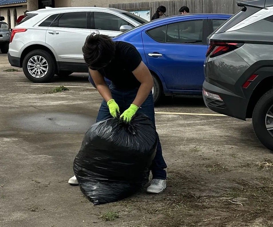 Person wearing gloves ties up a black trash bag in a parking lot. Several cars are visible in the background.