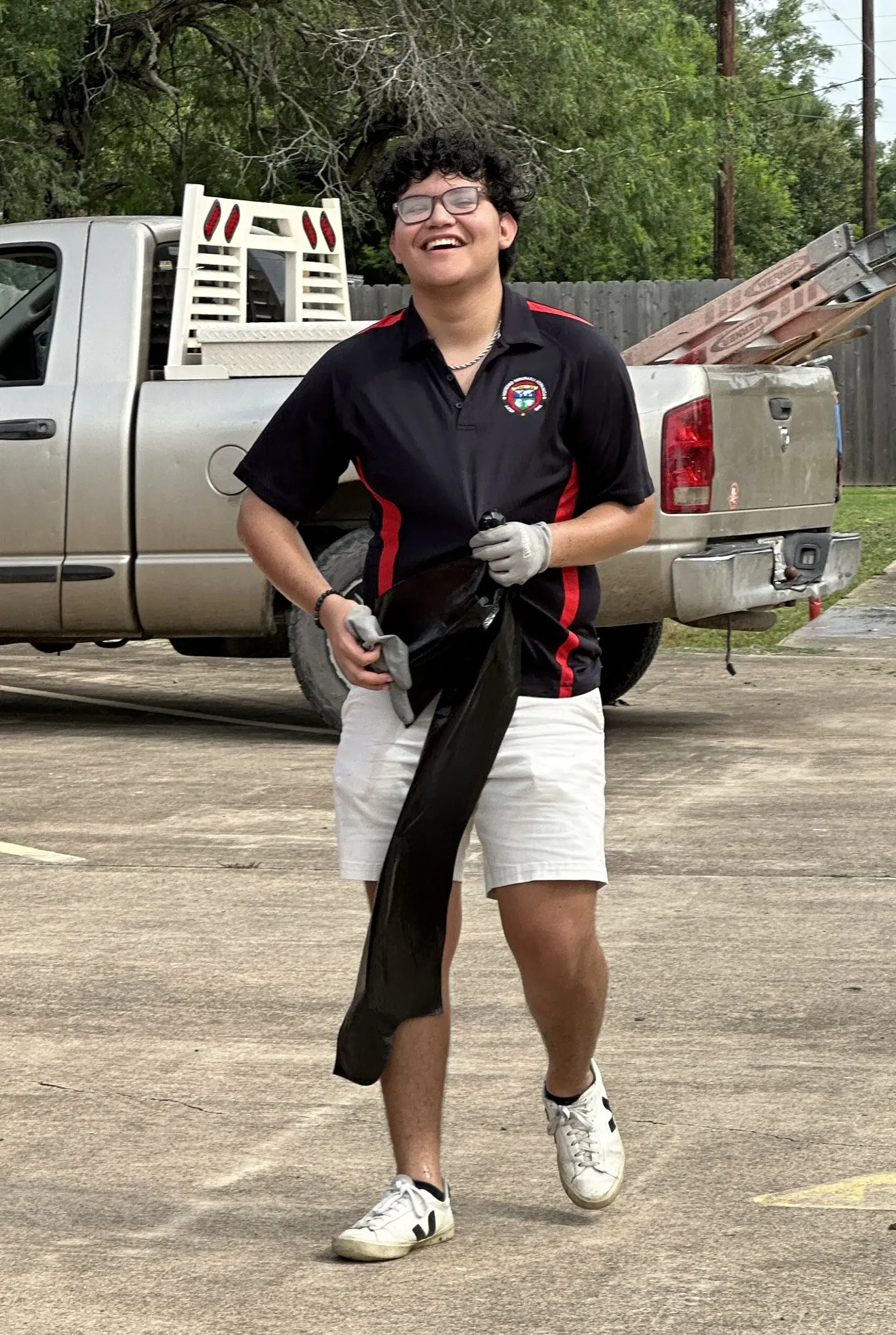 Young person in work attire smiles, holding a black item, with a truck in the background.