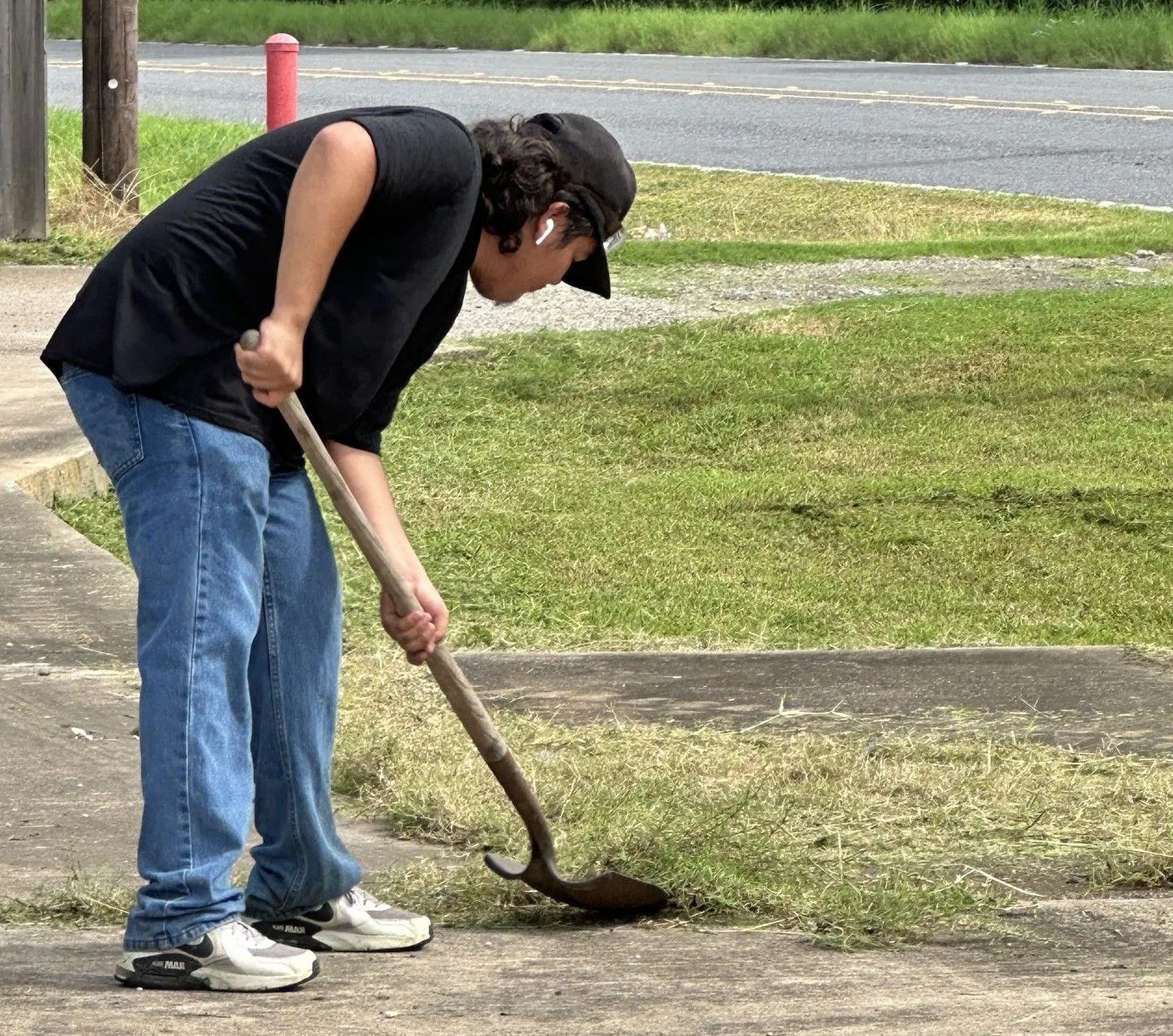 Person in black shirt and jeans uses a hoe to remove grass from a sidewalk.