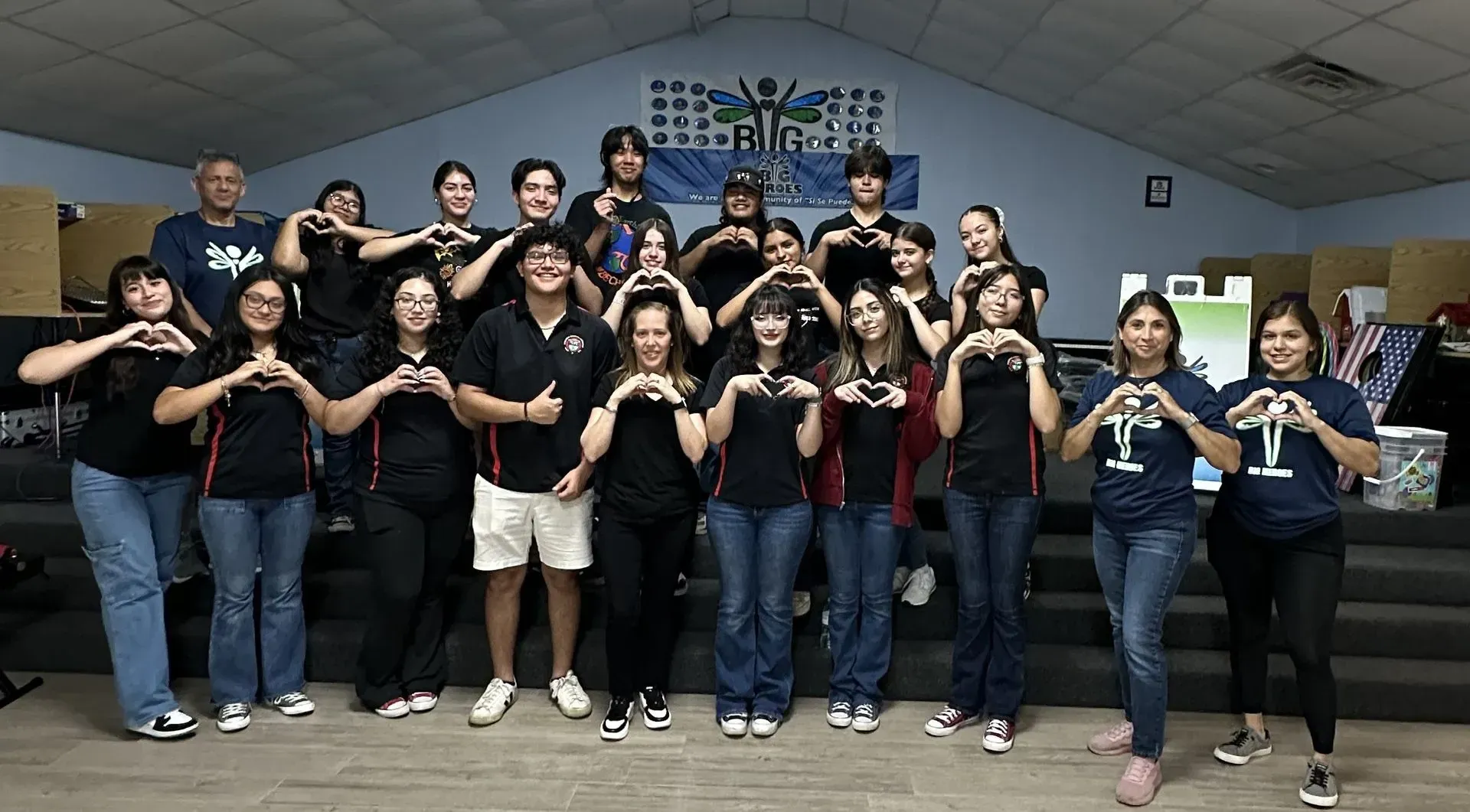 Group of people, mostly teens, indoors making heart shapes with their hands. Some wear black shirts.