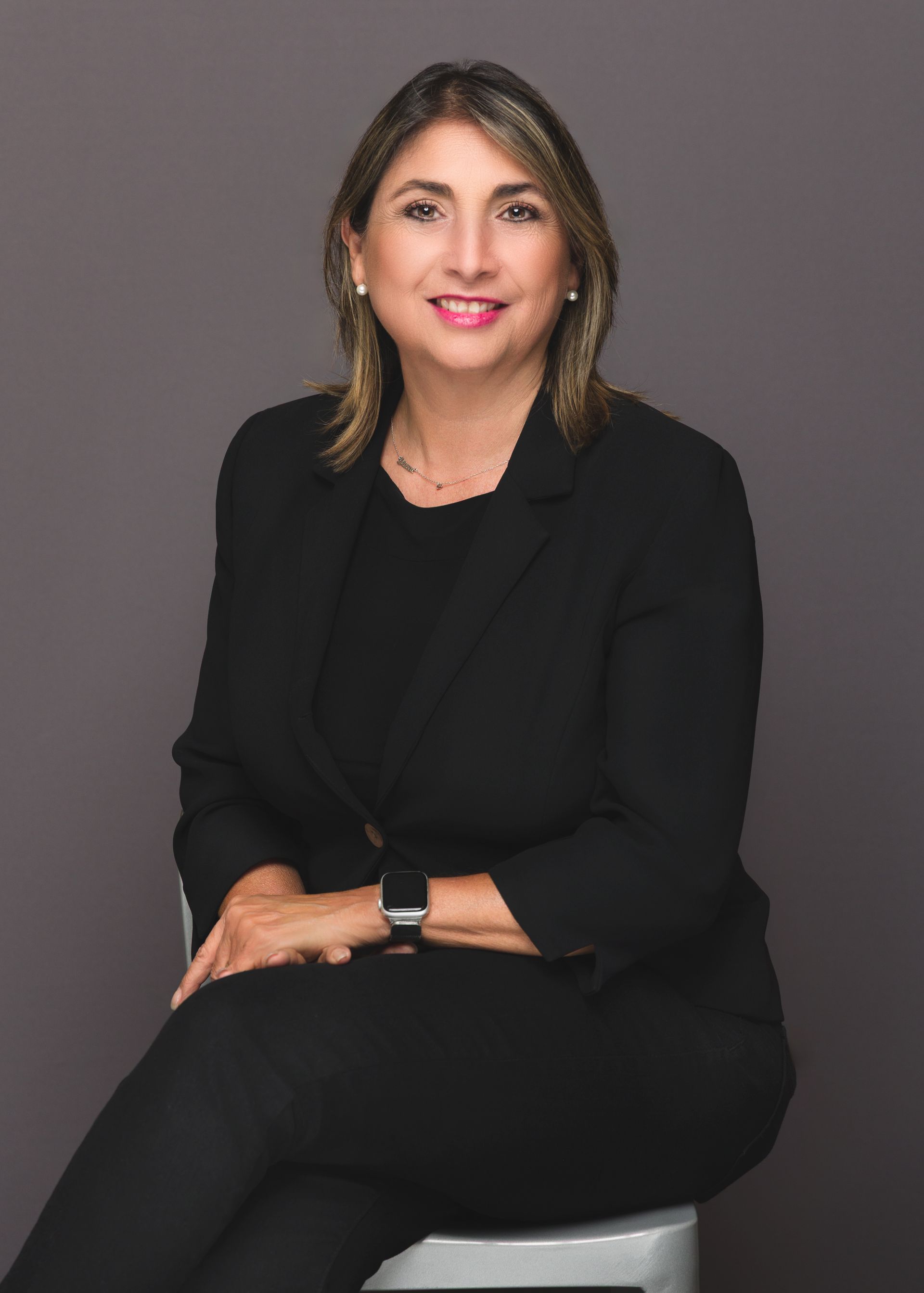 Woman in a black suit sits on a stool, smiling at the camera against a gray background.
