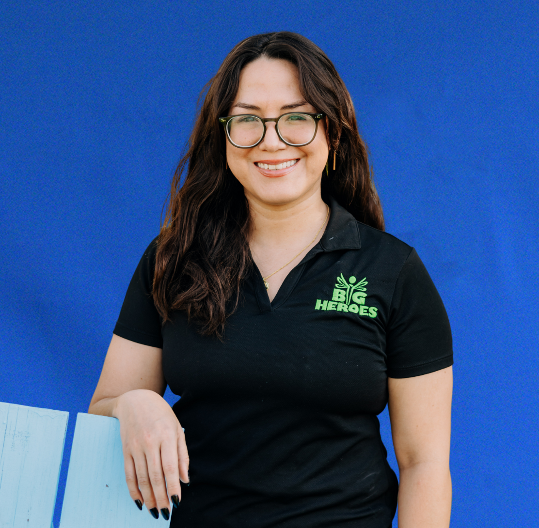 Woman with glasses in black shirt with “BIG HEROES” logo, leaning against blue wall.