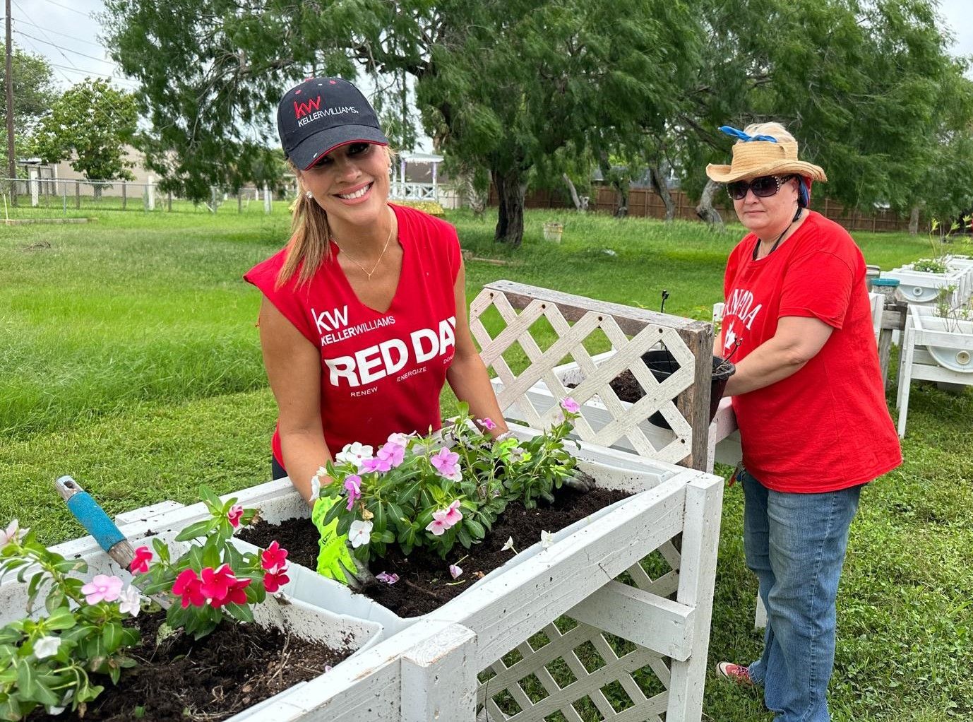 Two women in red shirts are planting flowers in a garden.