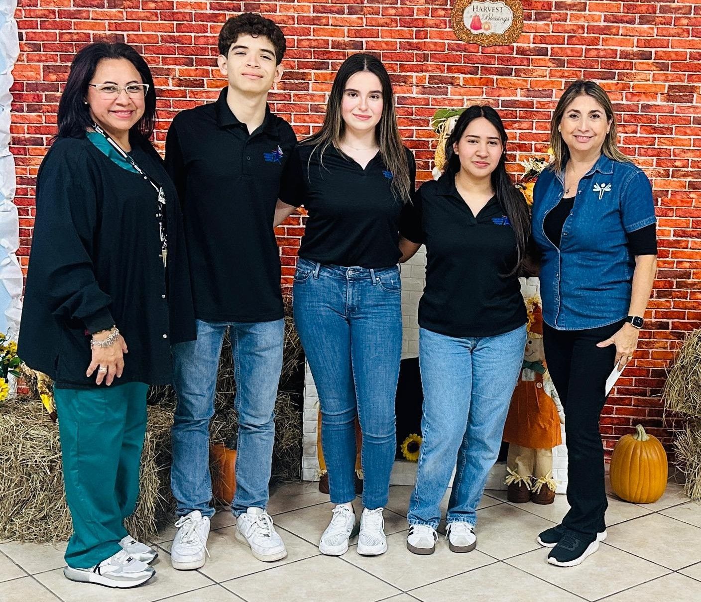 Five people smiling, posing for a photo against a brick wall. Four in black shirts and jeans, one in scrubs.