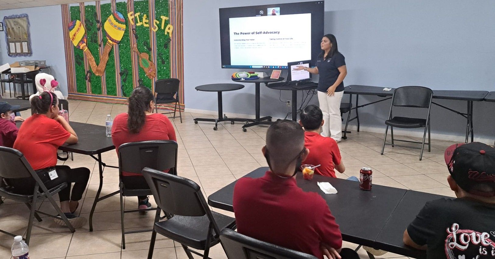 A woman presents to a group at tables in a community center. People wear red shirts.