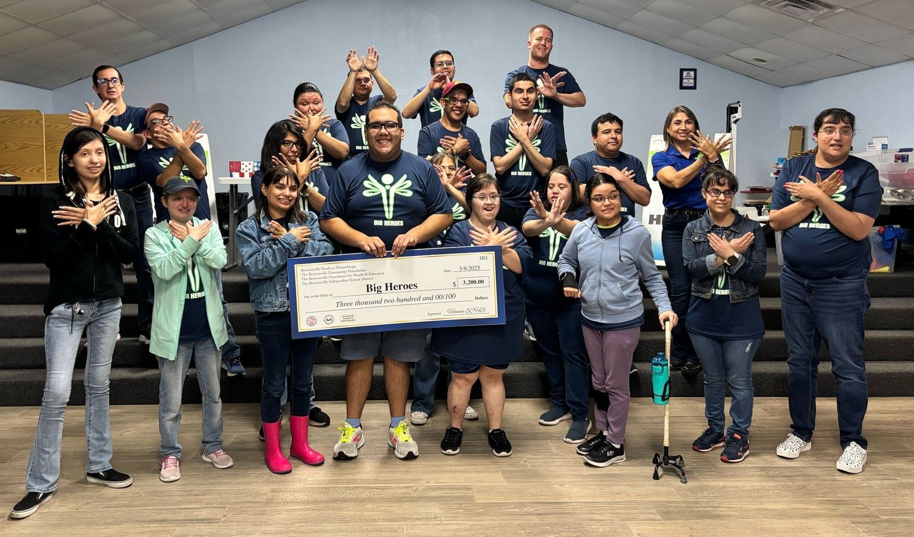 Group holding large check, posing with arms crossed. Blue shirts, mostly indoors.