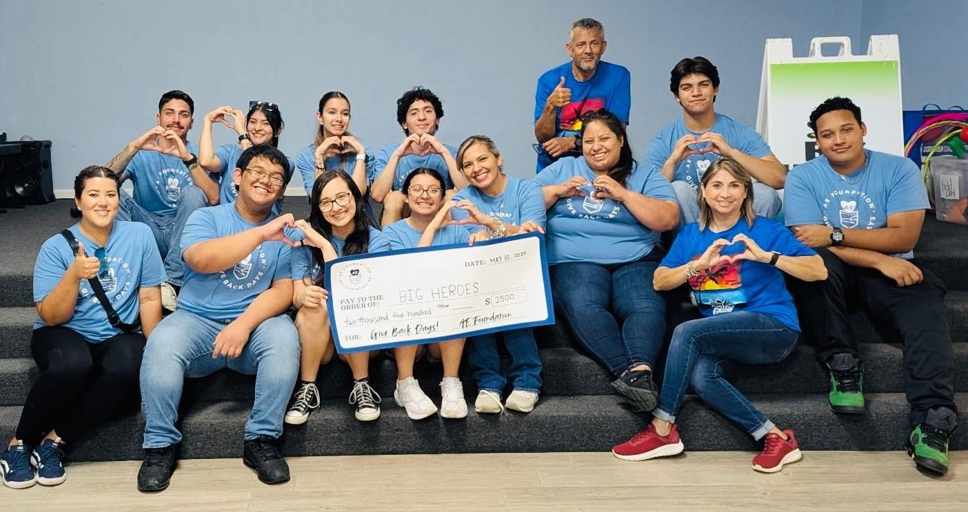 A group of people in blue shirts make heart shapes with their hands, posing with a large check.
