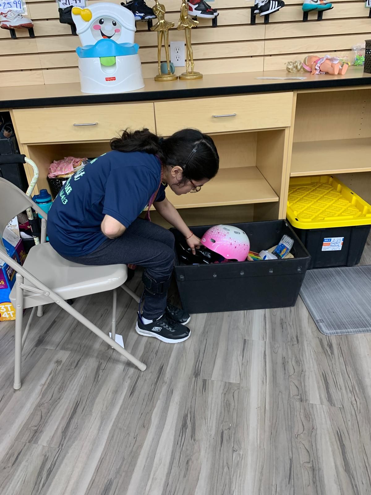 Woman looking in a bin, likely at a store, with items visible. She's wearing glasses and sitting on a chair.