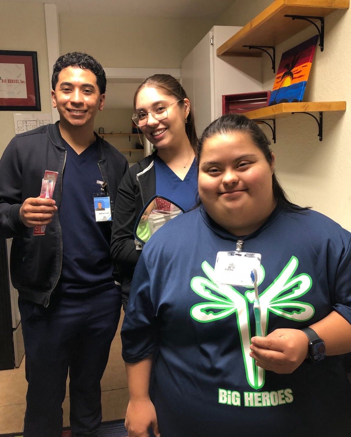 Three people smiling at camera, holding gifts. Indoors, near a shelf with art. One person has Down syndrome.