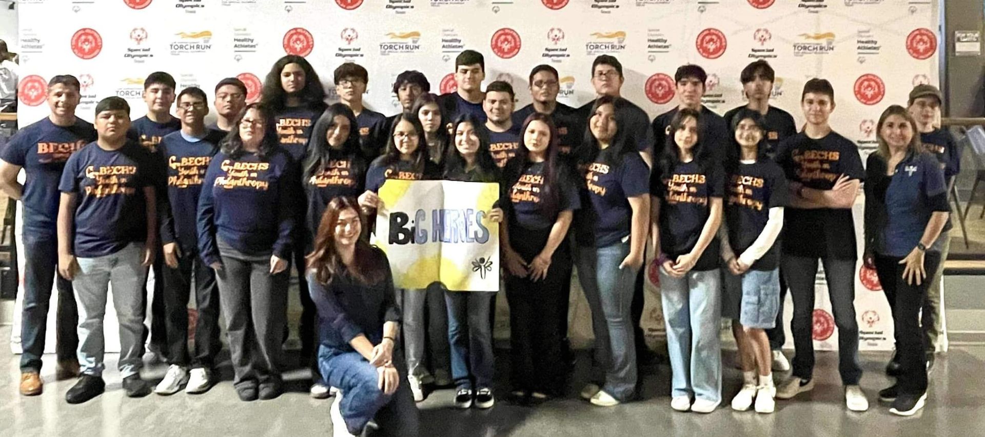 Group of students in blue shirts, holding a banner. They are posing in front of a patterned backdrop.