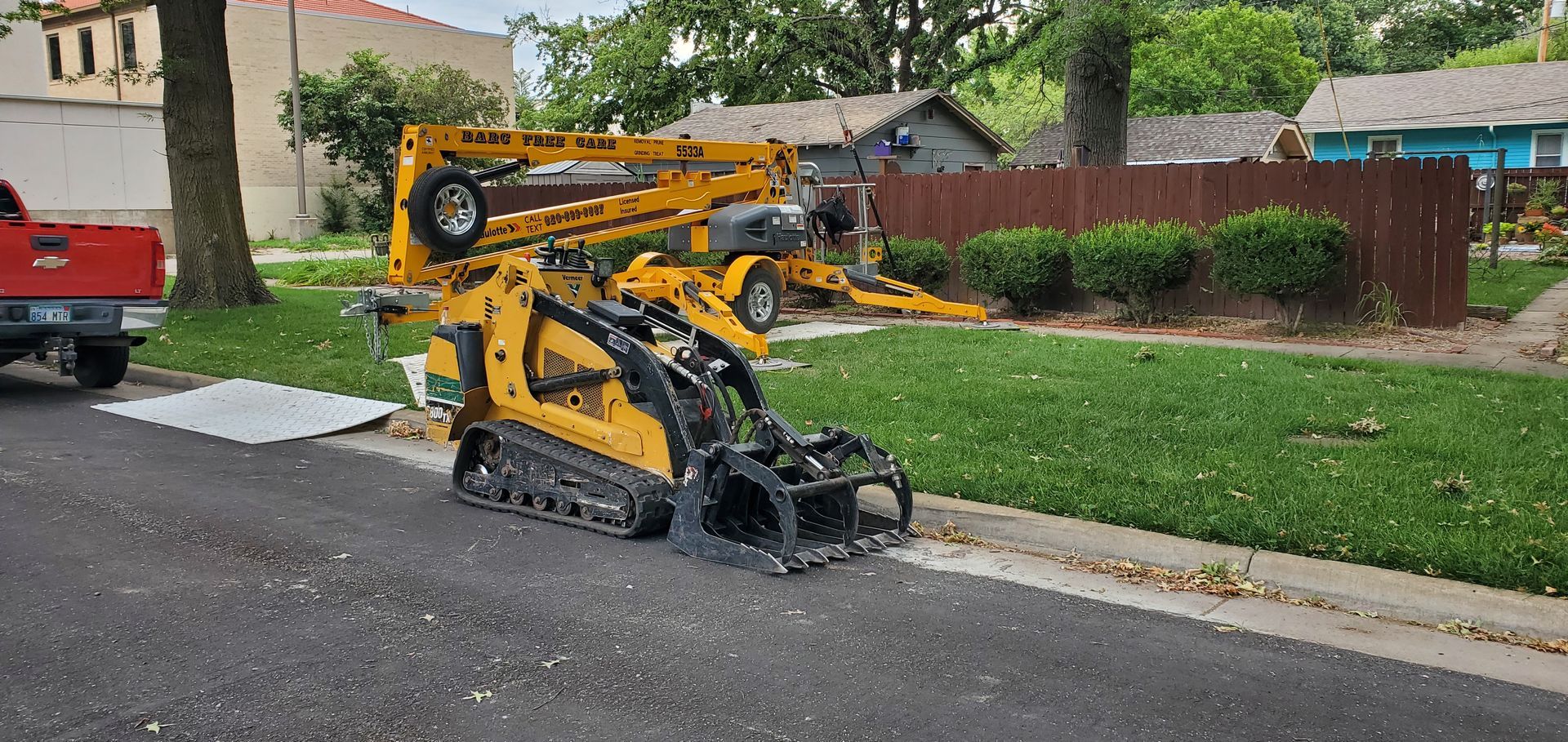 Yellow utility machine on a wet street beside a sidewalk and red truck, with a grass curb nearby
