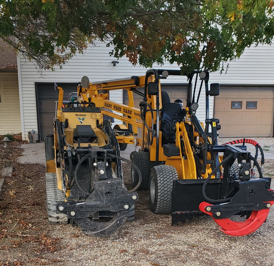 Yellow construction machines parked in a driveway under a tree, with a red attachment in front