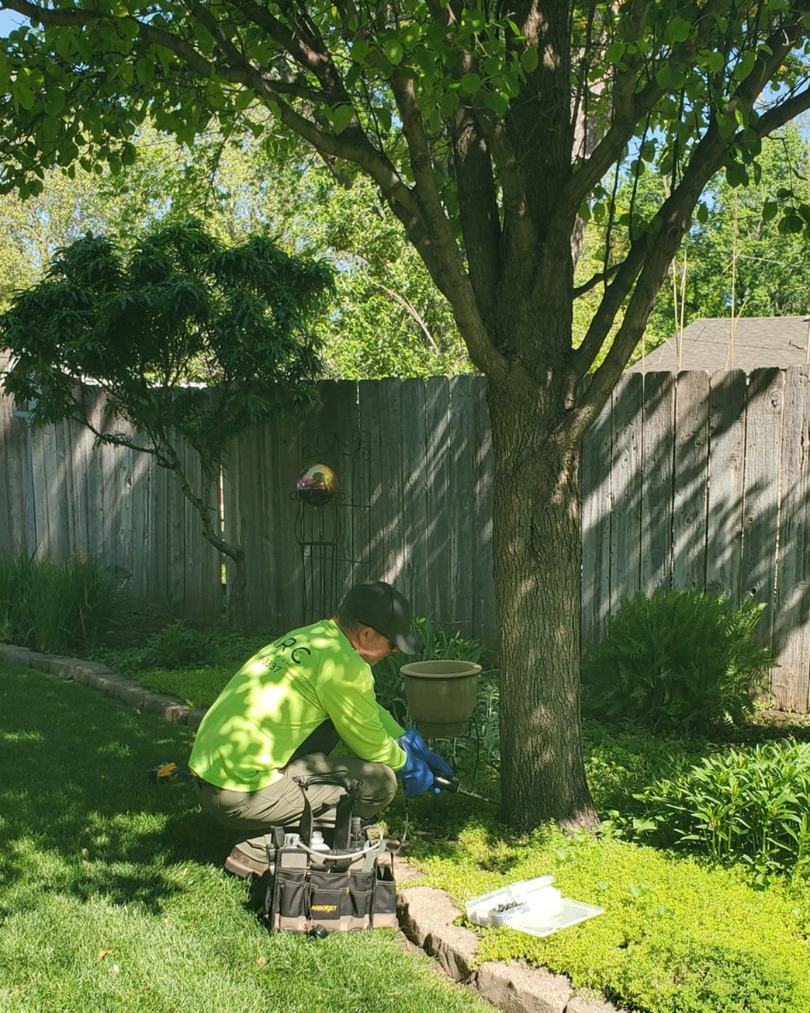 Person in a neon jacket working by a tree in a sunlit backyard with tools on the grass.