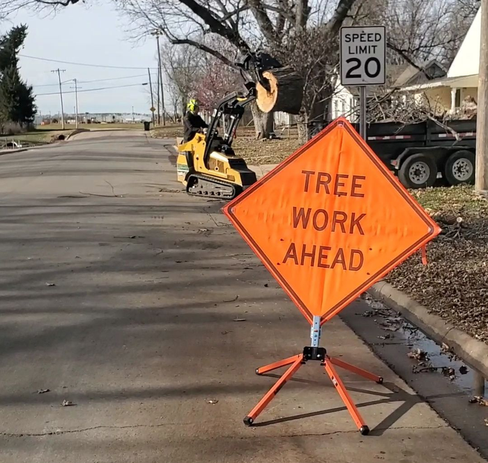 Orange “TREE WORK AHEAD” sign beside sidewalk with a speed limit 20 sign and tree trimming equipment nearby