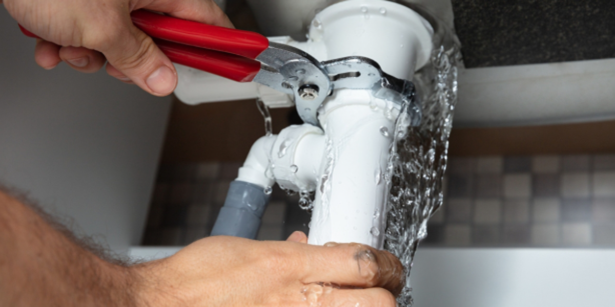 Plumber using pliers on a leaking white pipe under a sink; water is flowing out.