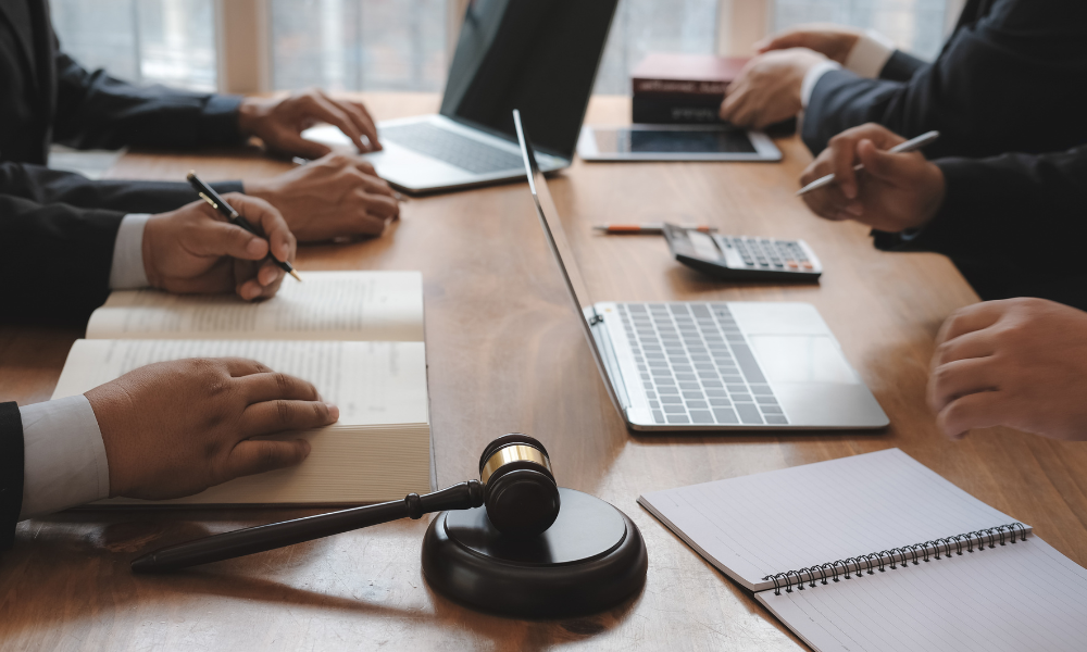 Legal professionals in suits at a table with books, laptops, a gavel, and notepad.