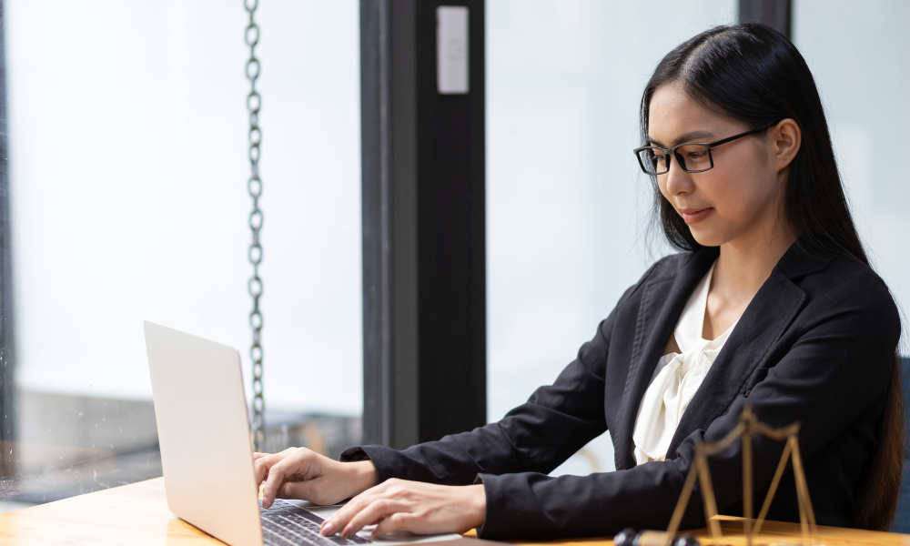 Woman in glasses and blazer working on laptop, next to window.