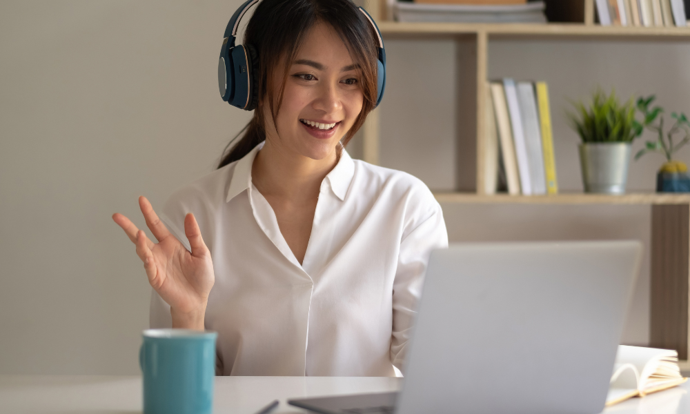 Woman wearing headphones waves while on a video call at a desk with a laptop and coffee mug.