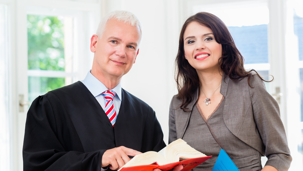 A judge in robe and a woman in a suit smiling, holding a book and papers, indoors.