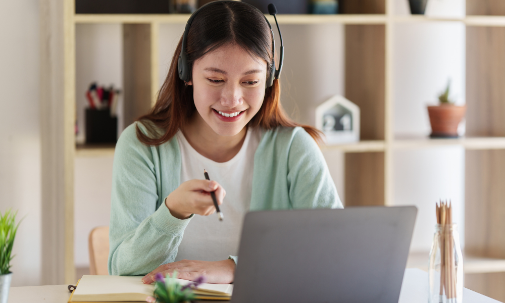 Woman wearing headset, smiling, using laptop at desk, holding pen. Light green cardigan.