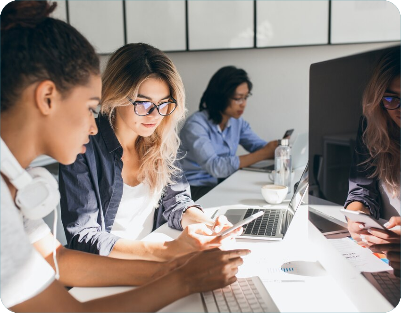 Four women working together at desks with computers and other devices in a modern office.