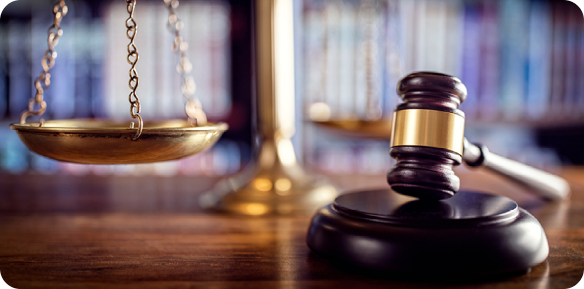 Scales of justice and gavel on a wooden table, in front of a blurred bookshelf.