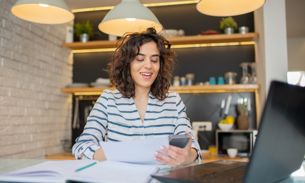 Woman smiling, holding phone and papers, working at a laptop in a kitchen with overhead lights.