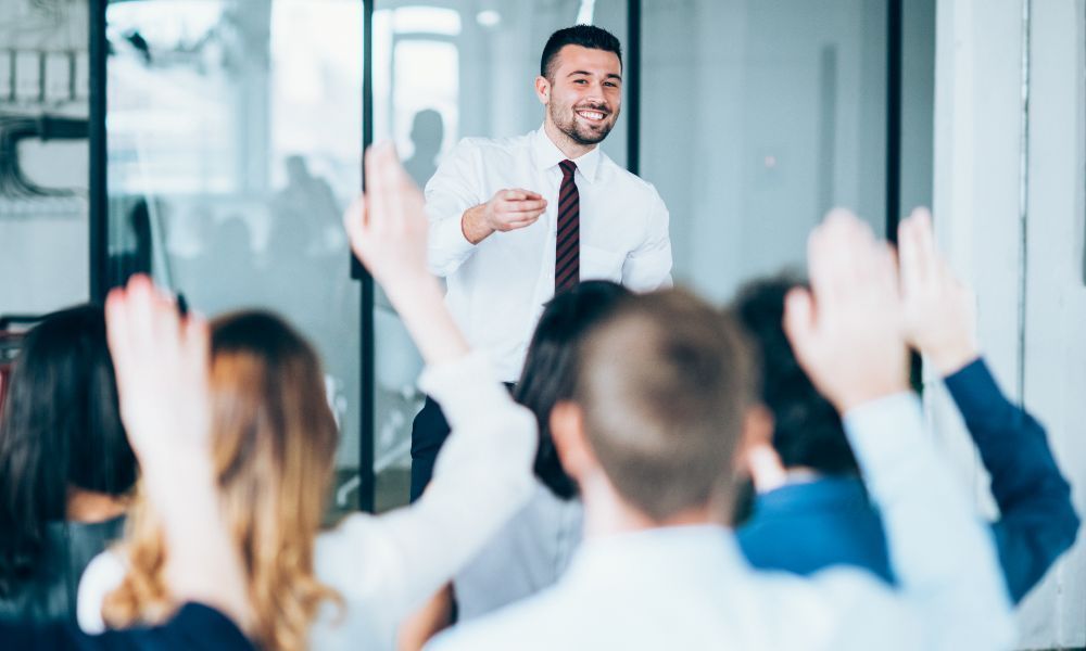 Man addressing a group with raised hands in a bright office.