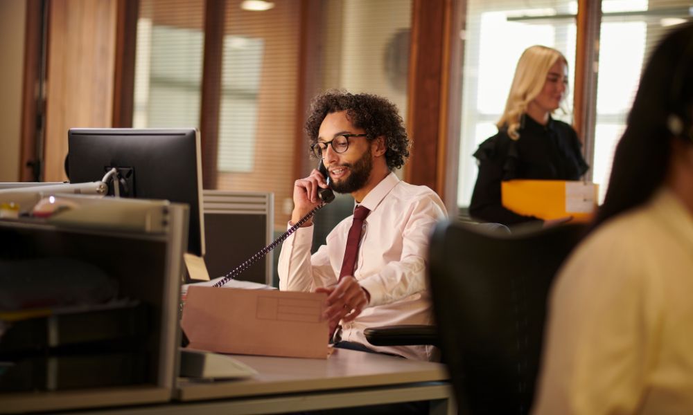 Man with glasses on phone at desk, holding papers; woman in background. Office setting.