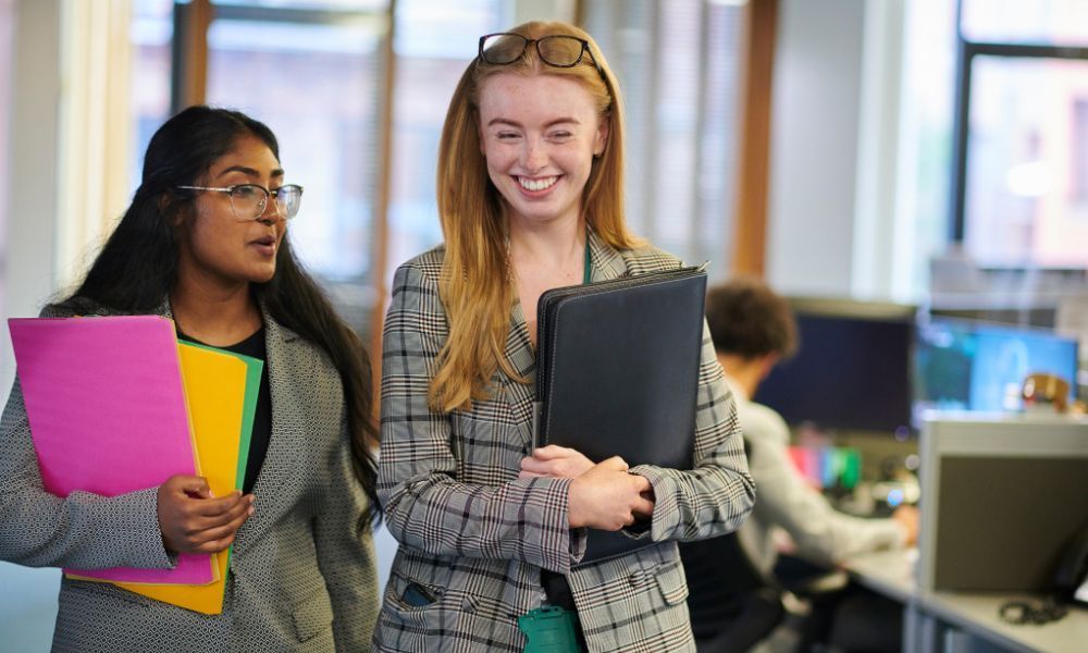 Two women in business attire walking in an office, one smiling and holding a folder, the other looking away and holding folders.