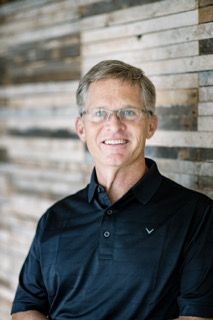 Connor wearing glasses and a black shirt is smiling in front of a wooden wall.