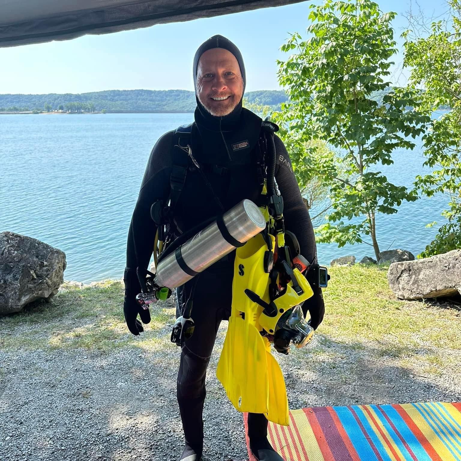 Kneale in a wet suit is holding a scuba tank and flippers.