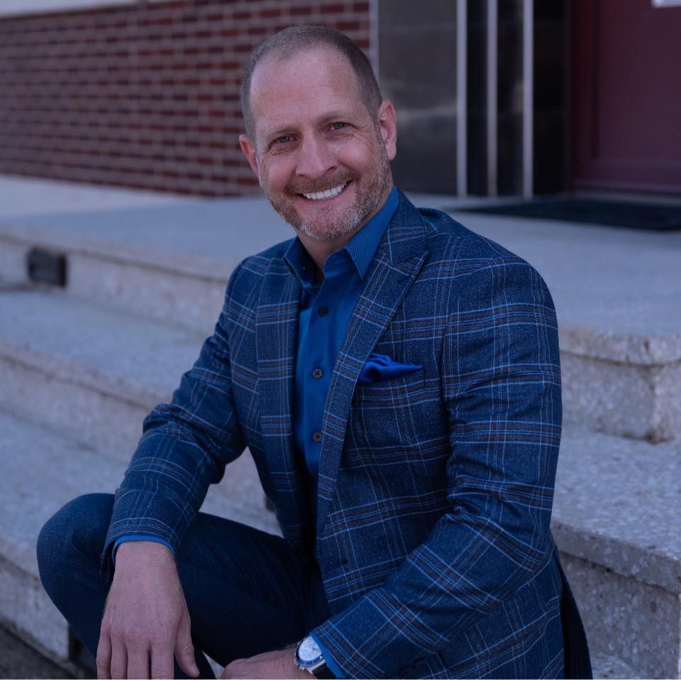 Kneale in a suit is sitting on the steps of a building and smiling.
