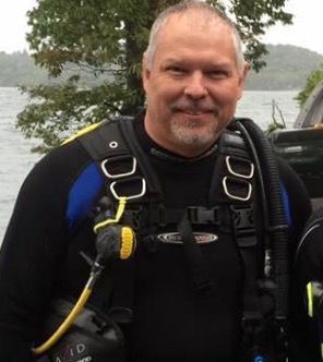 Roger wearing a scuba diving suit is standing in front of a body of water.