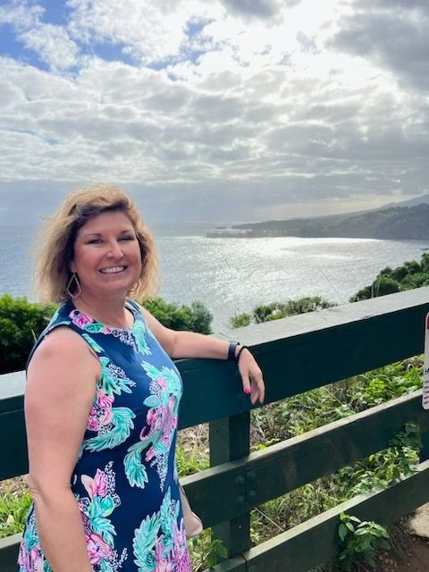 Shawna in a floral dress is leaning on a fence overlooking the ocean.