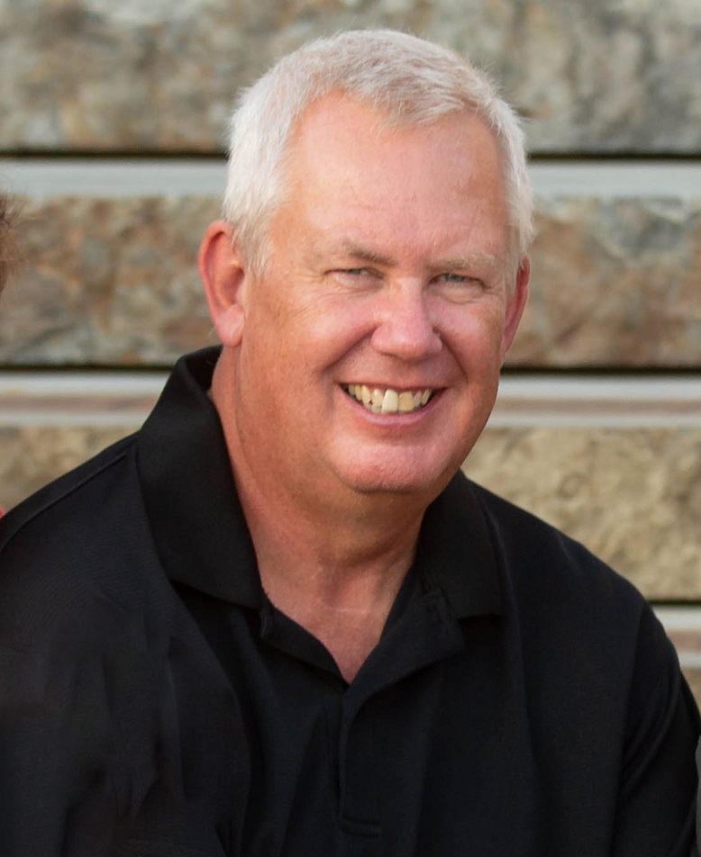 Walt in a black shirt is smiling in front of a stone wall.