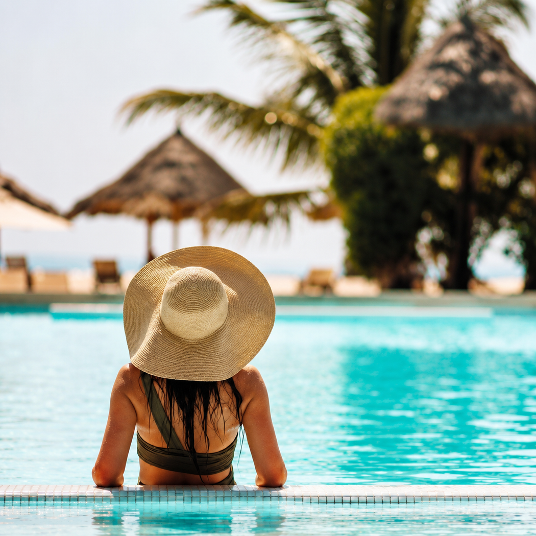 Una persona con un cappello di paglia si rilassa nella piscina turchese di un resort, con palme e cabine sulla spiaggia sullo sfondo.