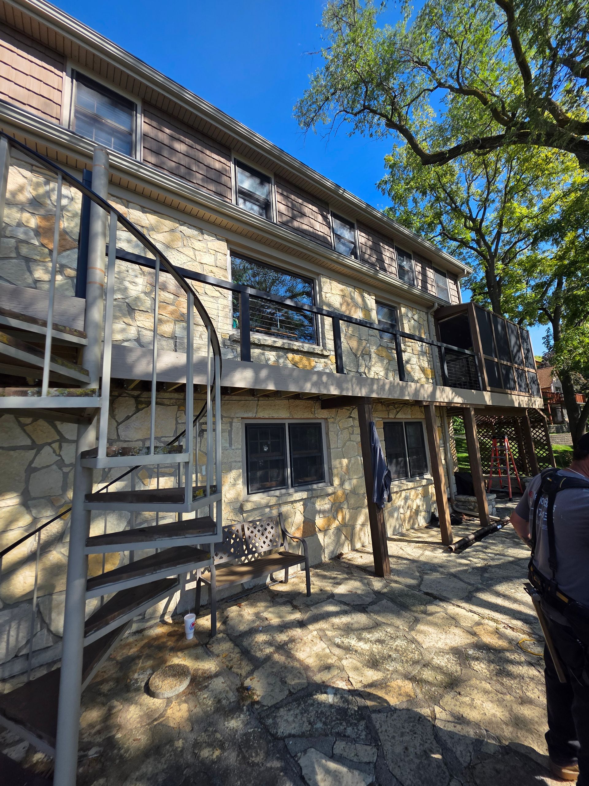 A stone house with a spiral staircase leading to a wooden deck, situated on a stone patio under a large tree - Danville, IL