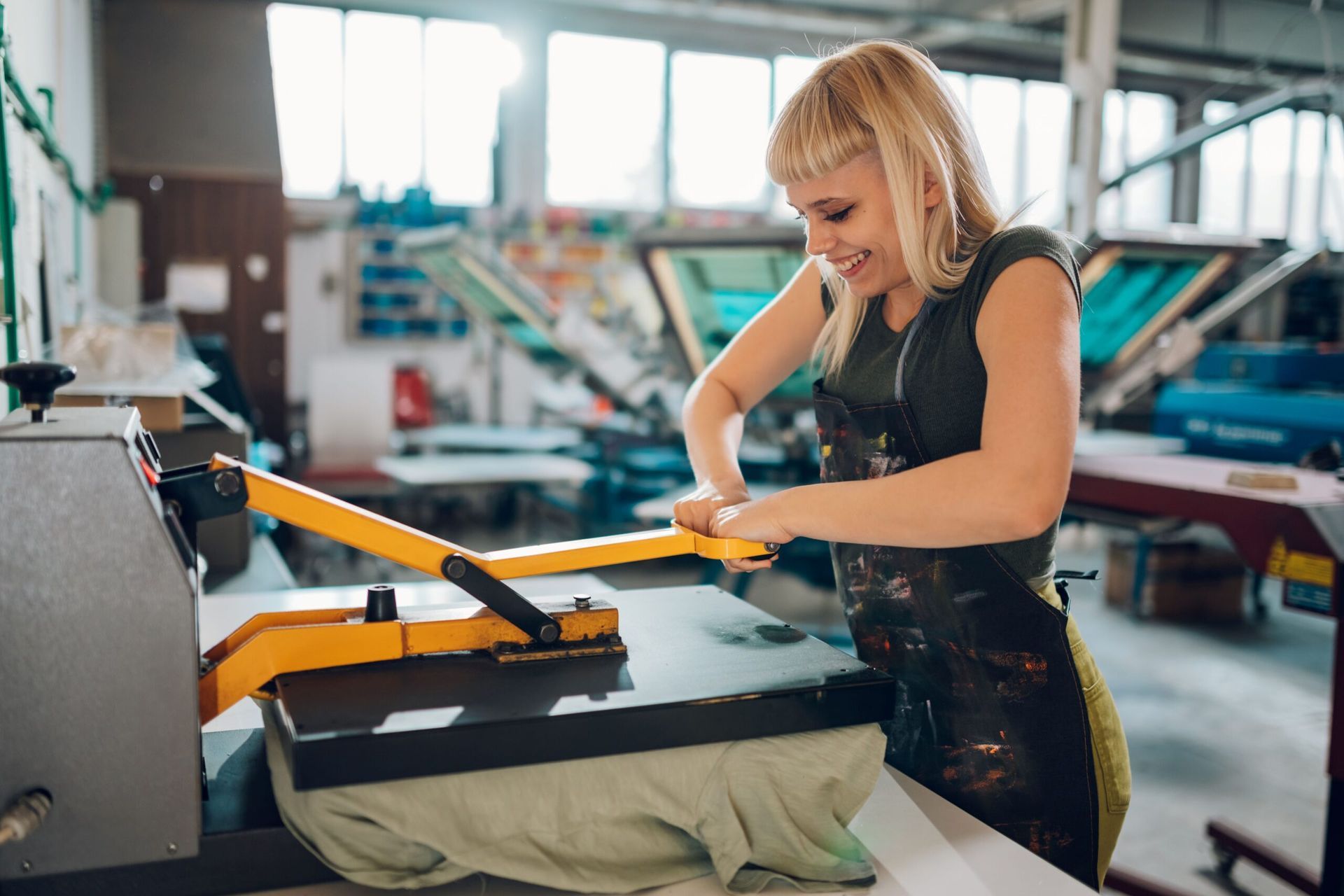 Woman in apron uses a heat press to print a design on a t-shirt at a print shop.