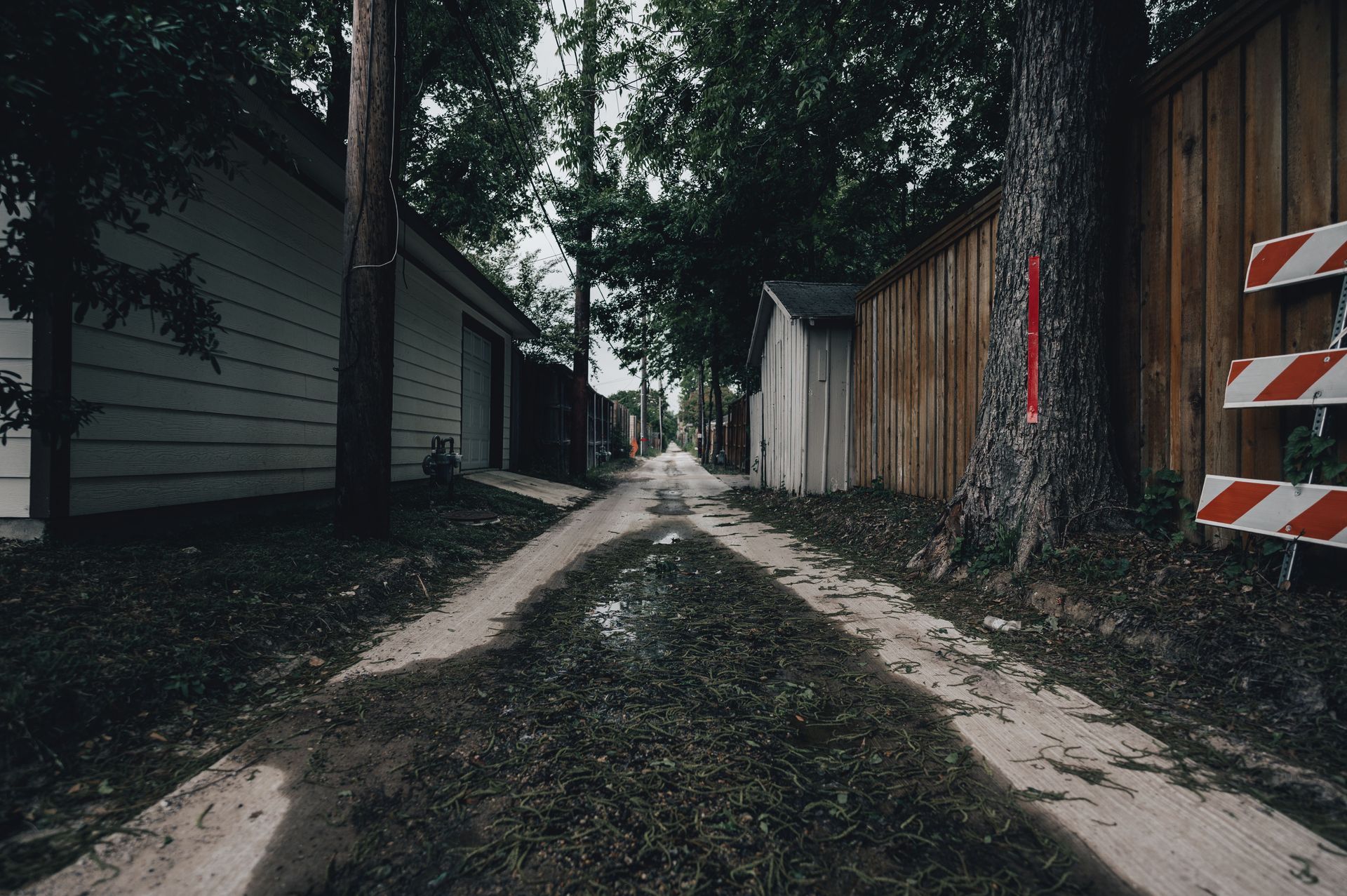 Narrow dirt path between buildings and trees, with a striped barrier on the right.