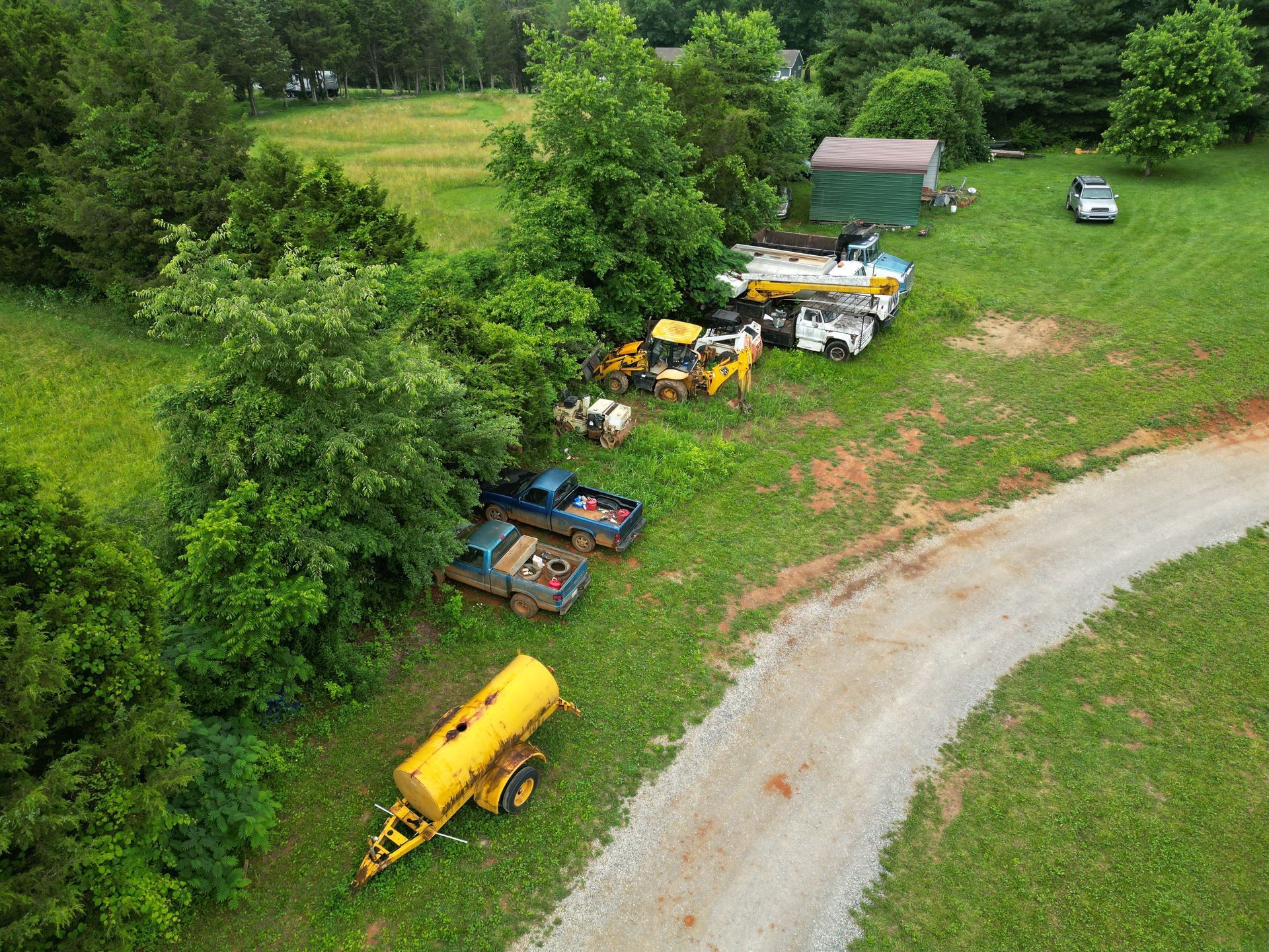 Aerial view of parked farm equipment and vehicles beside a gravel road and trees.
