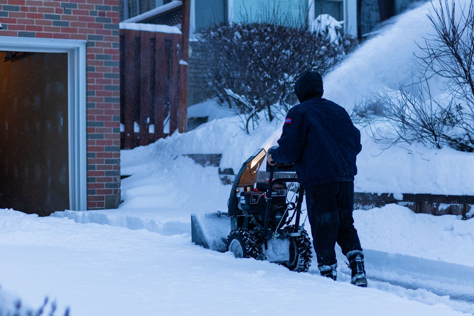 Person clearing snow with a snowblower beside a brick building and snow-covered path