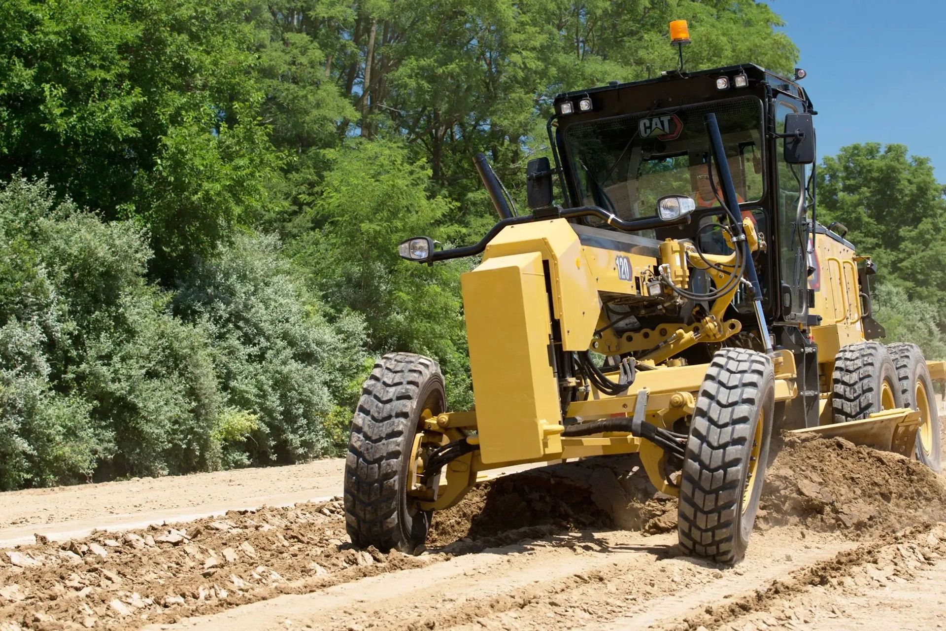 Yellow road grader working on a dirt road surrounded by green trees