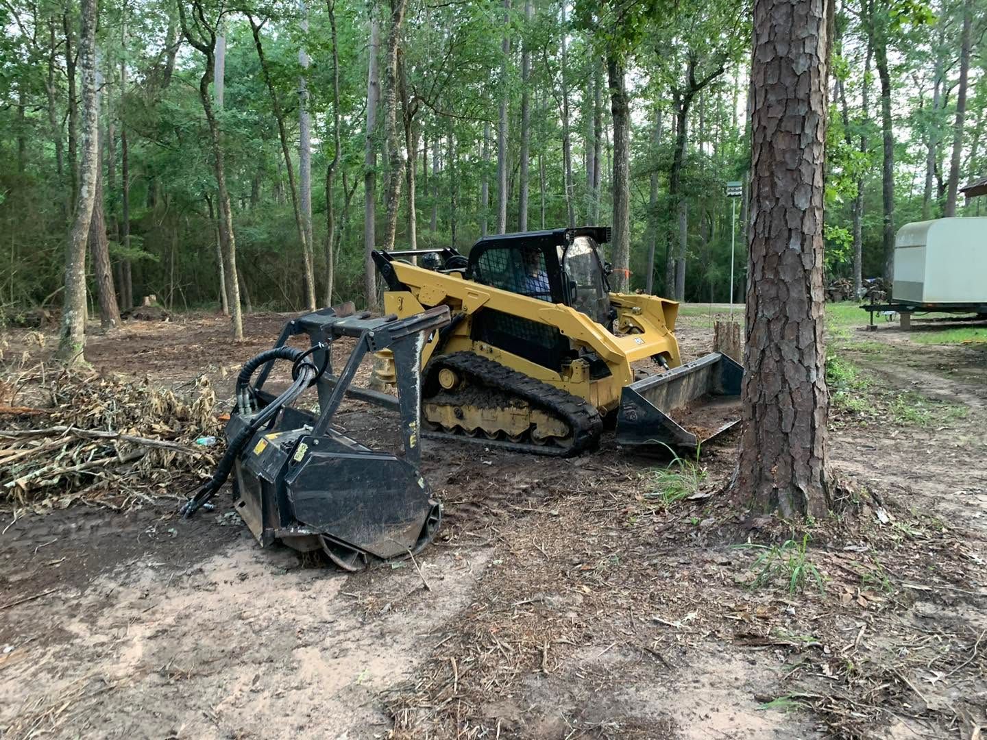 Yellow skid steer in a wooded area, pushing a black attachment beside a tree