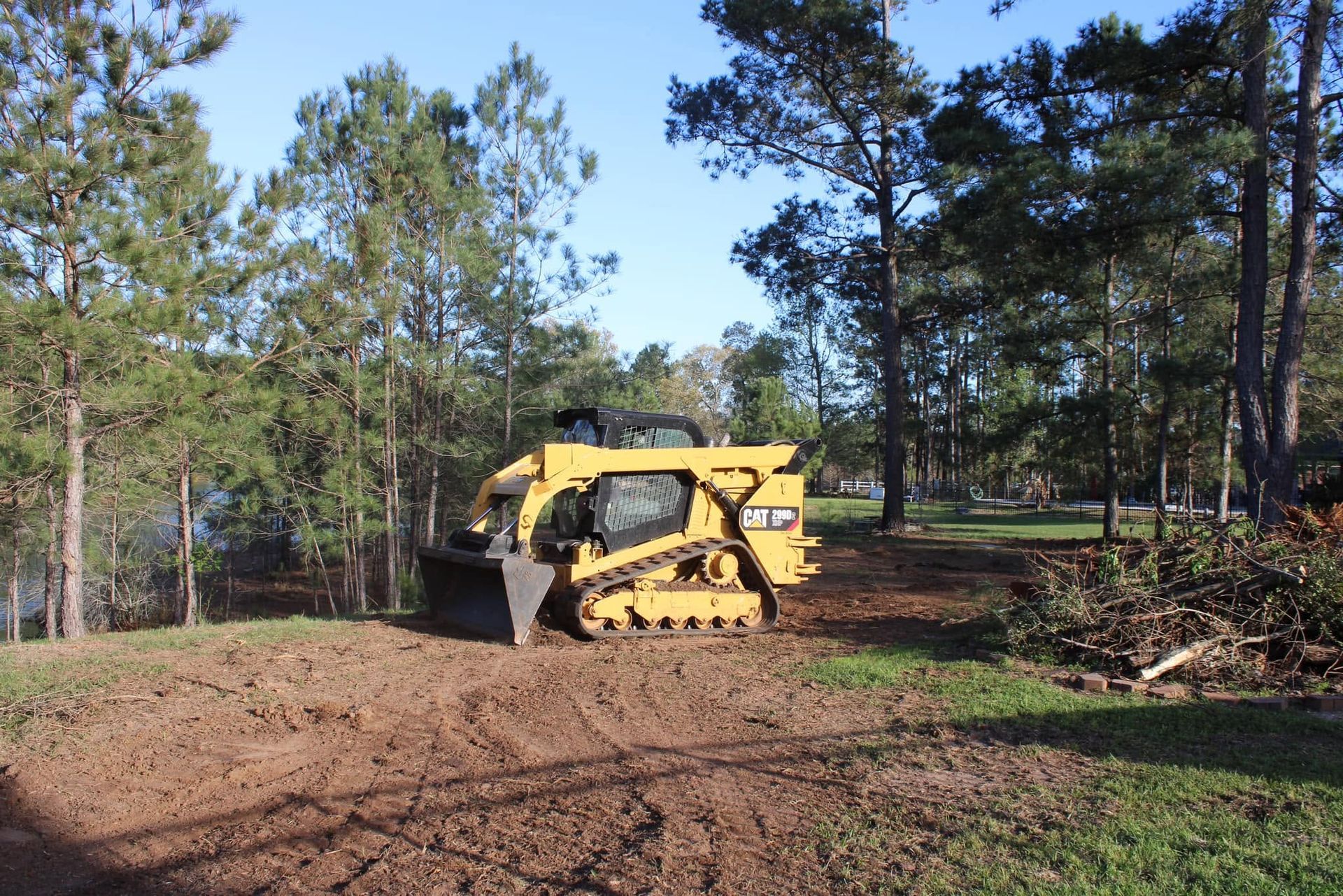 Yellow bulldozer clearing dirt on a wooded lot with pine trees and a pile of branches nearby