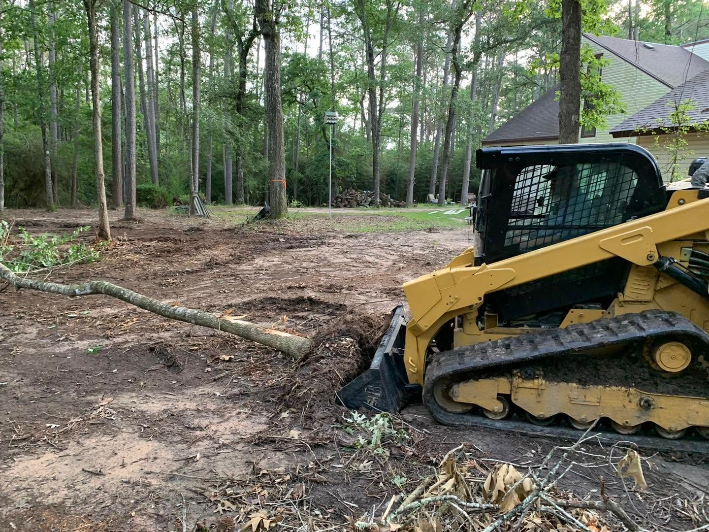 Yellow skid steer clearing dirt in a wooded area with a brush pile and freshly disturbed ground