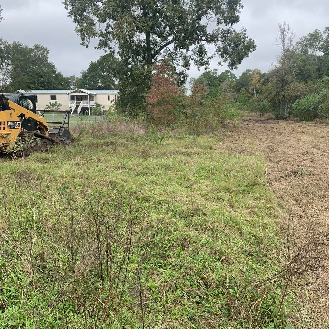 Overgrown field beside a house, with a yellow excavator clearing brush under cloudy skies