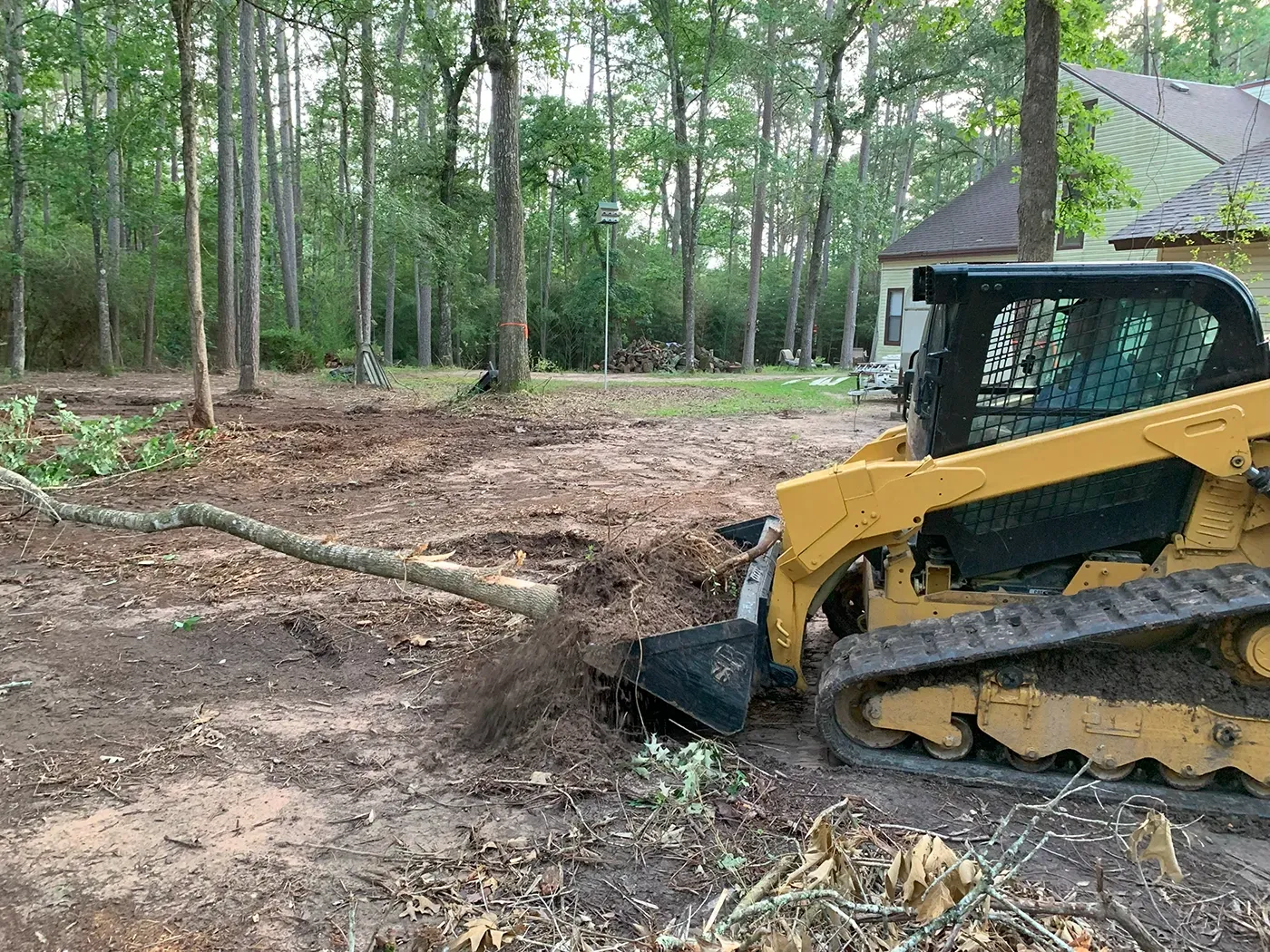 Yellow skid steer clearing dirt in a wooded area near a house.